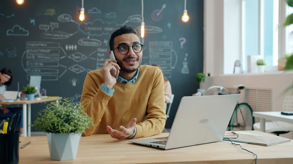 A man on a phone call at his desk, laptop open in front of him